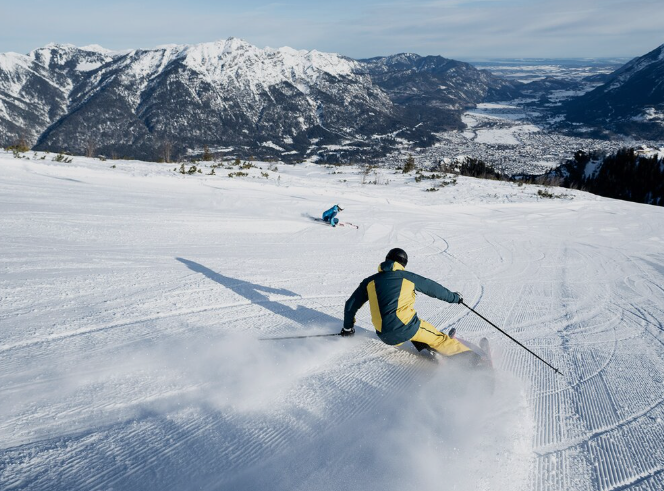 Ein Skifahrer carvt von hinten einen verschneiten Hang hinunter, wirbelt Schnee auf, mit einem weiten Gebirgstal im Hintergrund.