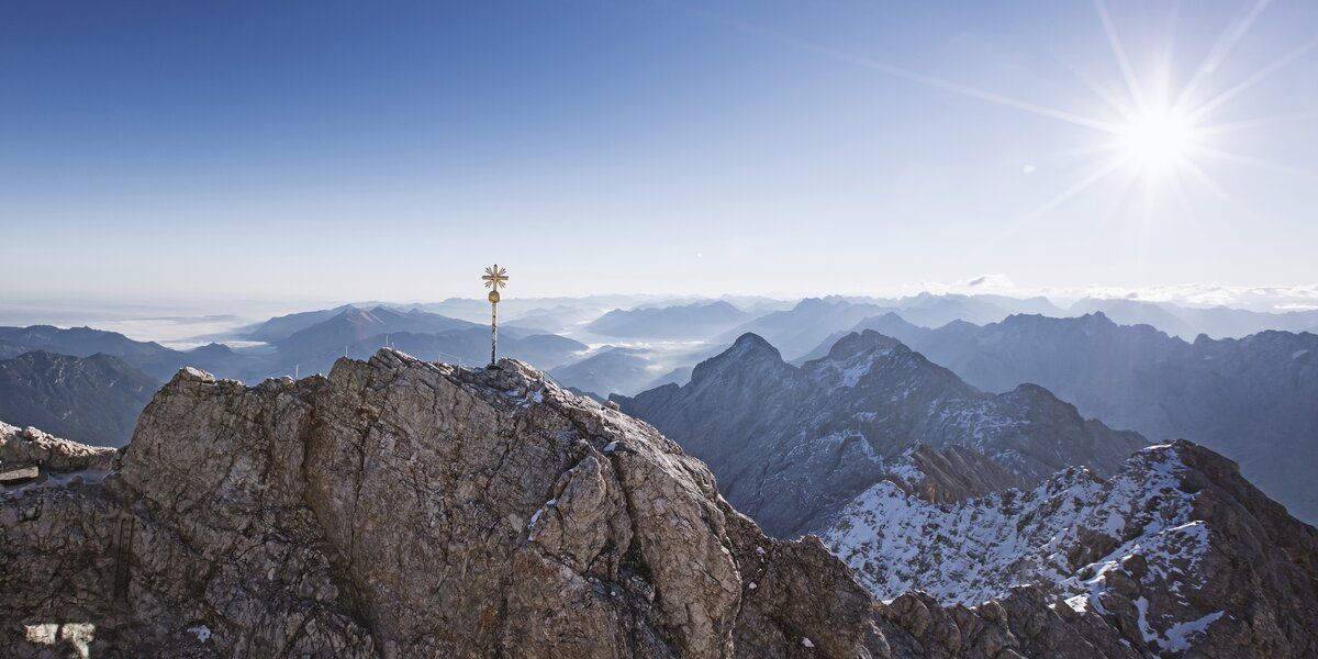 Ein goldenes Gipfelkreuz auf einem felsigen Gipfel überblickt eine weite Bergkette unter strahlender Sonne und klarem Himmel.