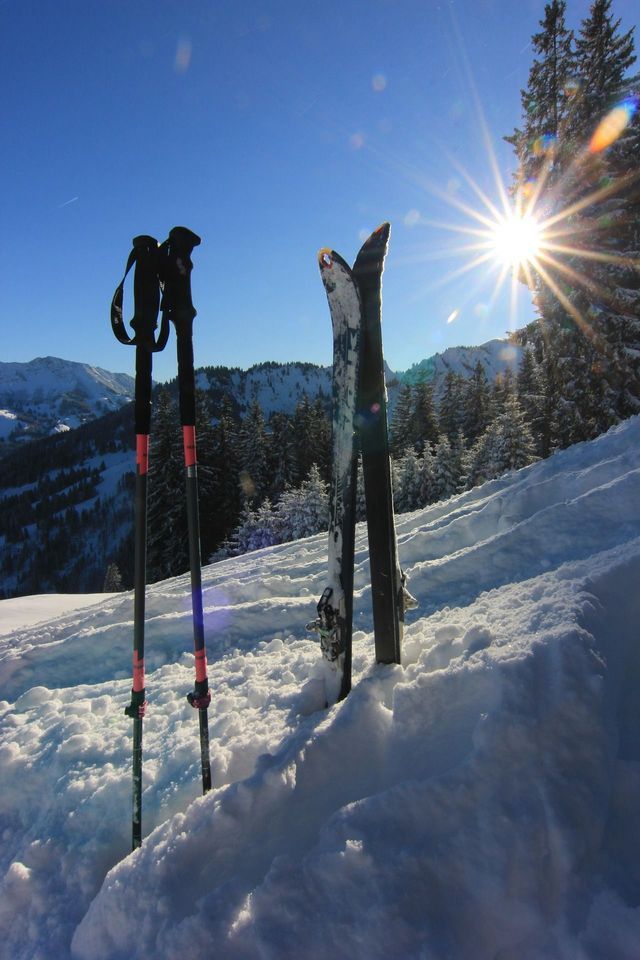 Ein Paar Ski und Skistöcke stecken aufrecht im Tiefschnee auf einem sonnigen Berg, mit einem hellen Sonnenstern am Bildrand.