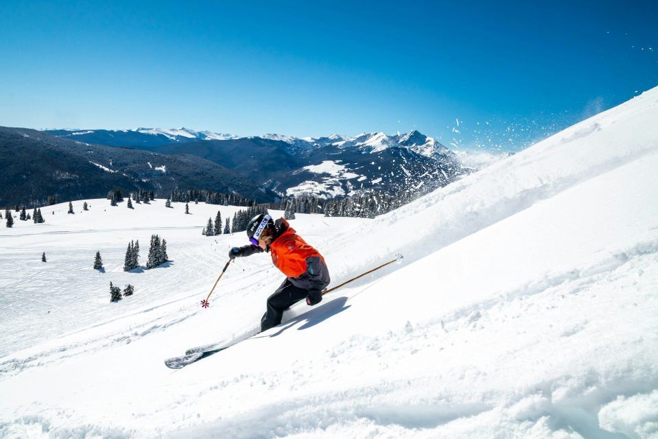 Ein Skifahrer in orangefarbener Jacke carvt einen Pulverschneehang hinunter, wirbelt dabei Schnee auf, unter klarem blauem Himmel und mit fernen Bergen.