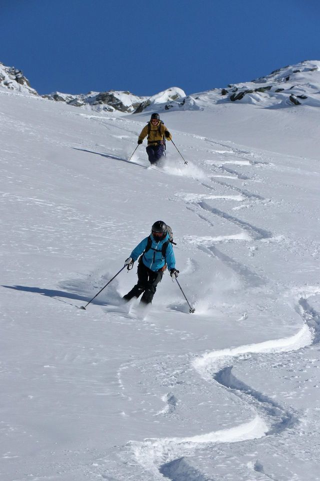Zwei Personen einer WeRoad Gruppenreise skifahren einen sonnigen, schneebedeckten Berg hinunter und hinterlassen geschwungene Spuren im Pulverschnee.