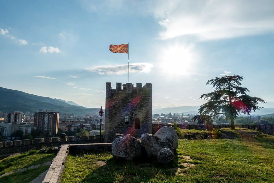 Die Flagge Nordmazedoniens weht von einem steinernen Burgturm auf einem Hügel, mit Blick auf eine Stadt und Berge unter strahlender Sonne.