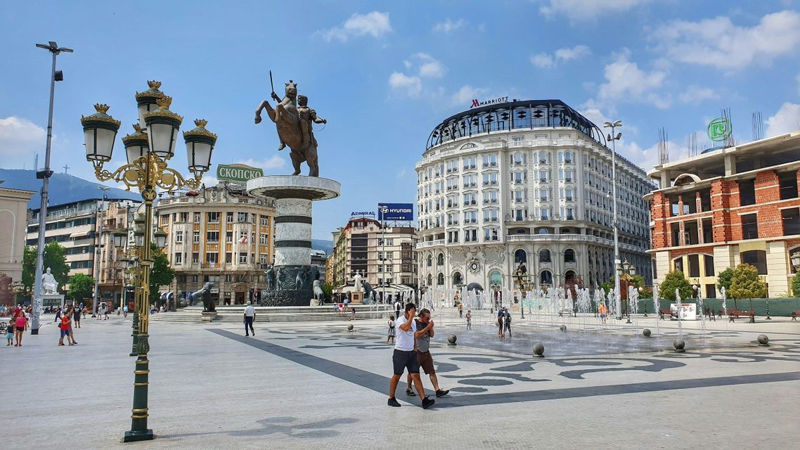 Eine große Reiterstatue steht auf einem Stadtplatz mit Springbrunnen, umgeben von Gebäuden und Menschen unter blauem Himmel.