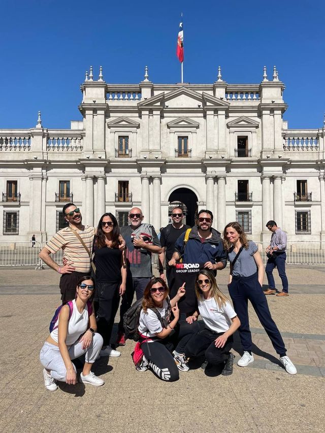 Un gruppo WeRoad in viaggio sorride per una foto in una piazza soleggiata, davanti a un imponente edificio classico con una bandiera.