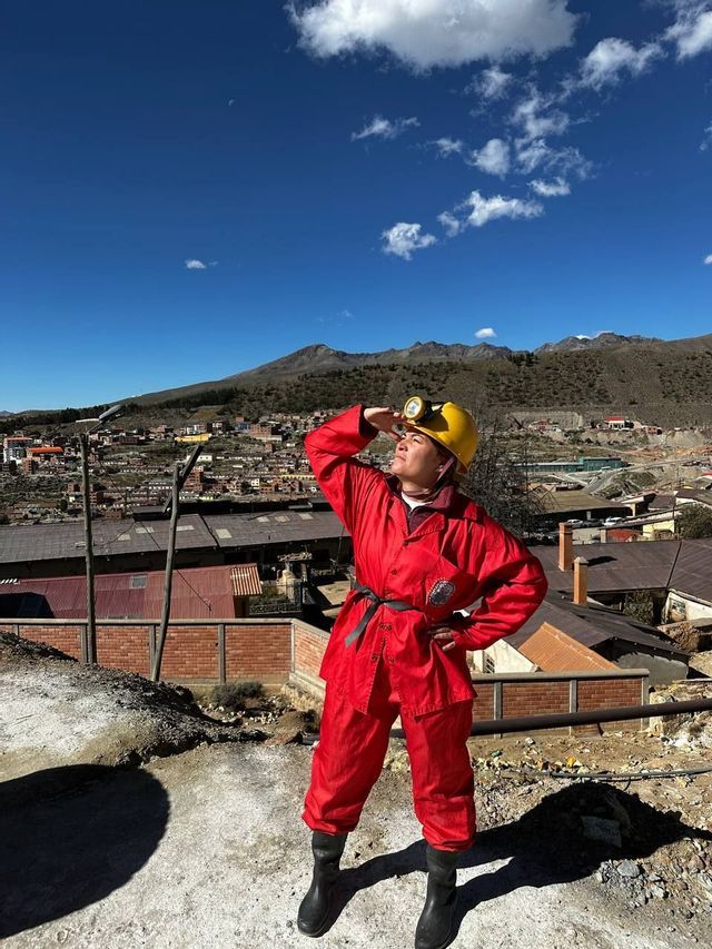 A woman wearing a red protective suit and a yellow hard hat with a headlamp stands on a hill overlooking a mining town.