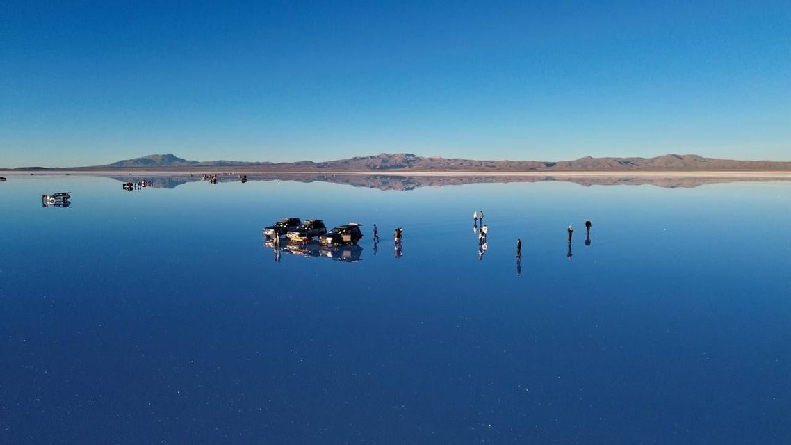 A WeRoad group trip stands on a vast, reflective salt flat with 4x4 vehicles, creating a perfect mirror of the blue sky and mountains.