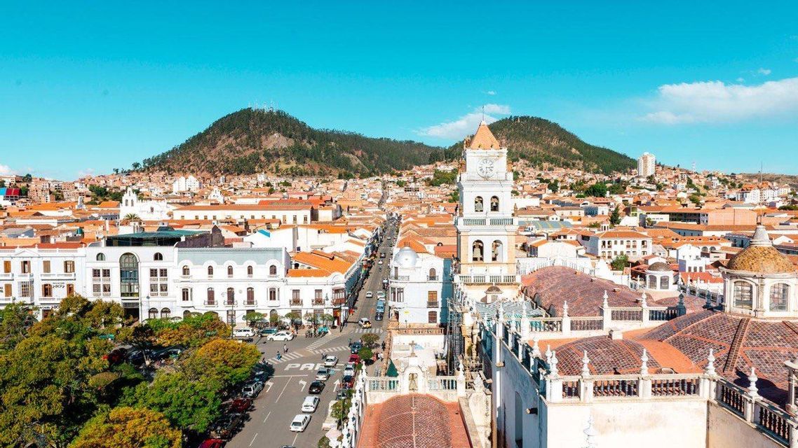 An elevated view of a cityscape with a white bell tower, orange tiled roofs, and green hills under a clear blue sky.