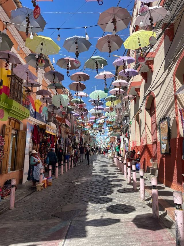Colorful umbrellas hang over a busy cobblestone market street, casting distinct shadows on the ground as people walk between shops.