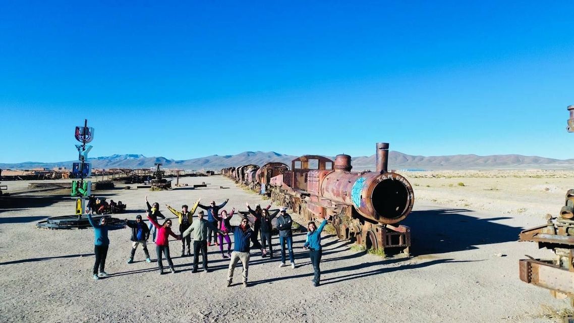 Un gruppo WeRoad si mette in posa con le braccia alzate davanti a un lungo treno arrugginito e abbandonato in un paesaggio desertico.