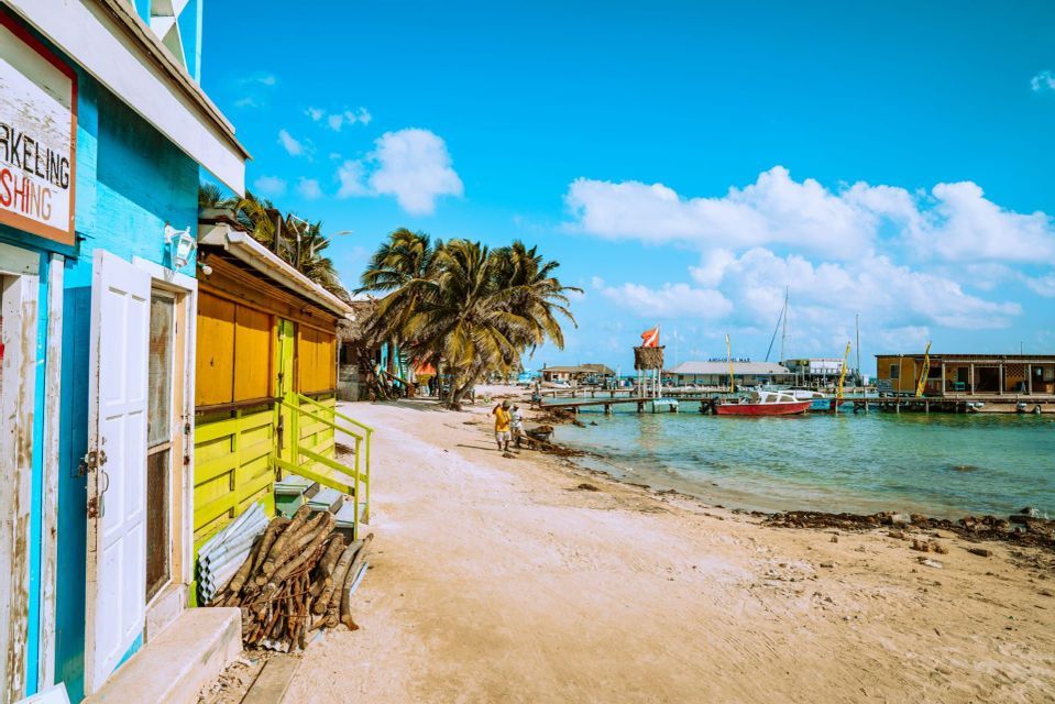 Bunte Holzgebäude und Palmen säumen einen Sandstrand neben einem Pier mit Booten auf türkisfarbenem Wasser.