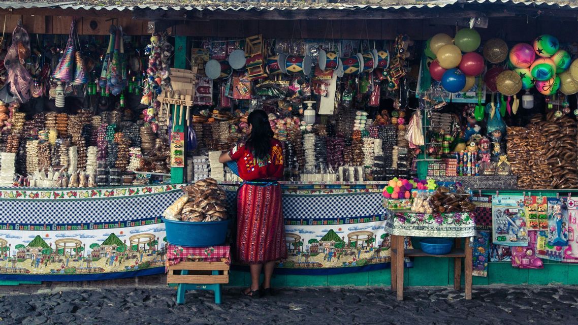Eine Frau in traditioneller Kleidung steht mit dem Rücken zur Kamera an einem farbenfrohen Marktstand, gefüllt mit Süßigkeiten und Spielzeug.