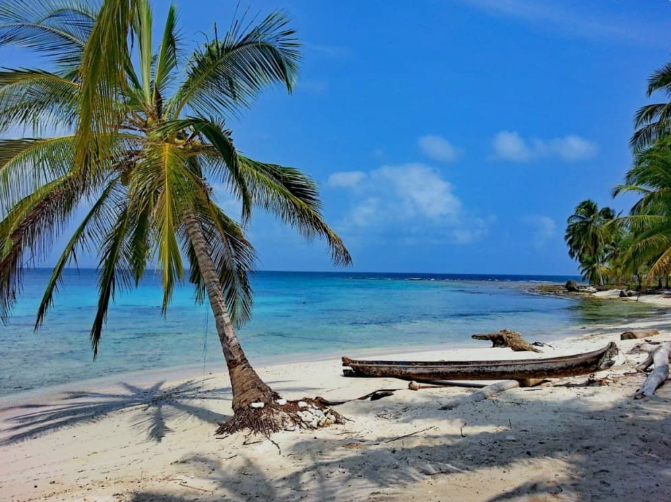 Eine Palme neigt sich über einen weißen Sandstrand mit einem Holzkanu, neben ruhigem türkisfarbenem Wasser unter blauem Himmel.