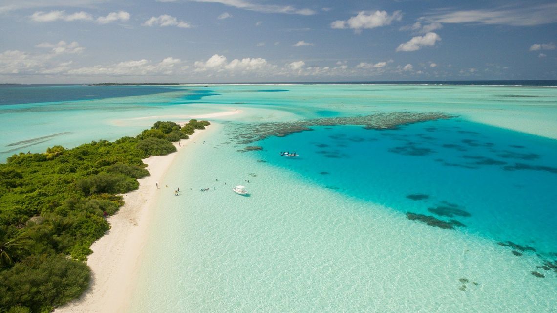 Una vista aerea di un banco di sabbia tropicale con vegetazione lussureggiante, circondato da acqua turchese dove le persone nuotano e le barche sono ancorate.