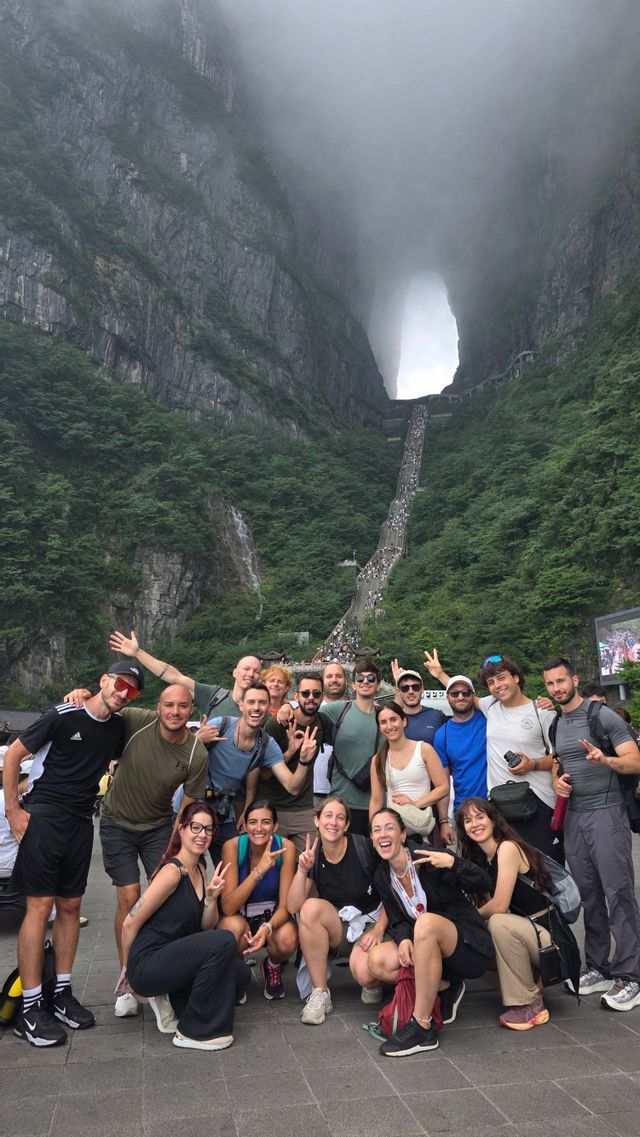 Un gruppo WeRoad in viaggio si fa fotografare ai piedi di una montagna ripida e verde, con un grande arco naturale e una lunga scalinata.