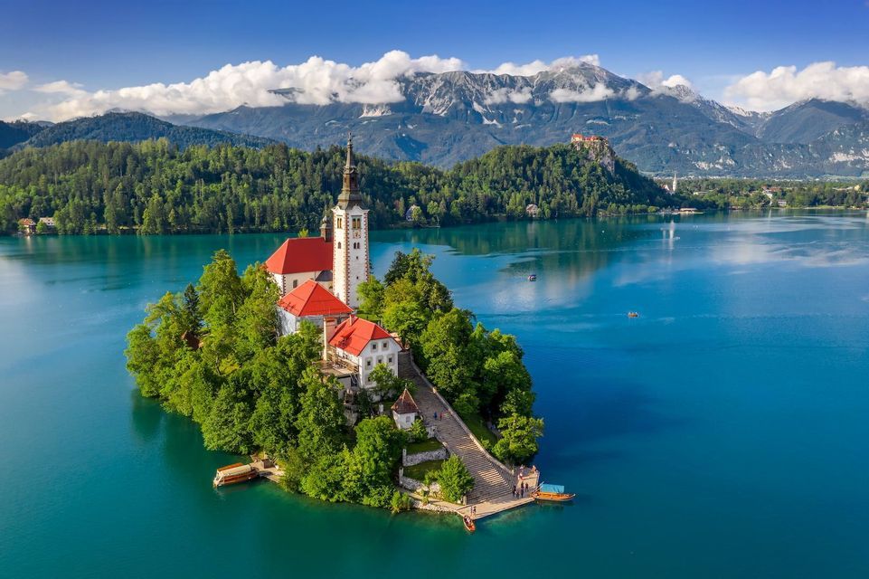 A white church with red roofs sits on a tree-covered island in a turquoise lake, with steep mountains in the background.