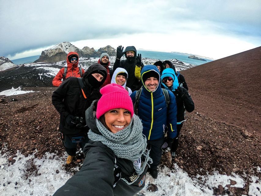 Un grupo de WeRoad, equipado para el invierno, se toma una selfie en una colina rocosa y nevada, con montañas y el mar de fondo.