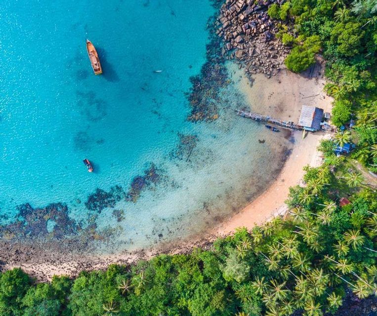 An aerial view of a tropical bay with clear turquoise water, two boats, and a sandy beach with a wooden pier and palm trees.