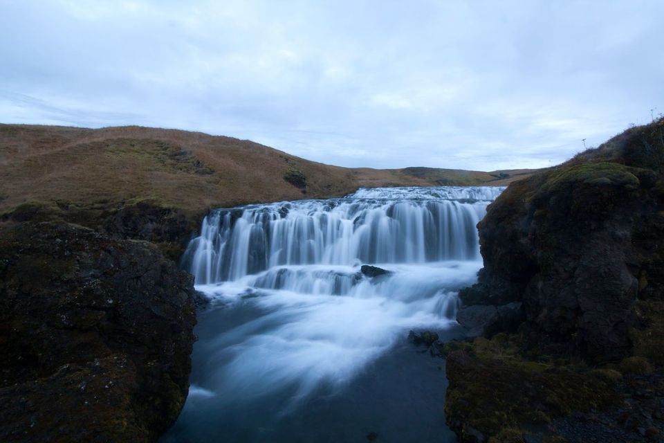 Una amplia cascada capturada con exposición prolongada, que cae sobre rocas entre dos oscuros acantilados y verdes colinas.