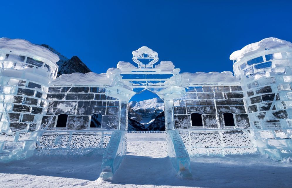 Un castello fatto di grandi blocchi di ghiaccio con un arco centrale si erge nella neve, con le montagne visibili sullo sfondo contro un cielo blu.