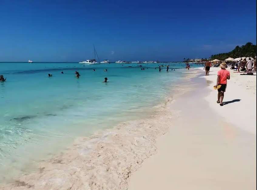 Une journée ensoleillée sur une plage tropicale avec des gens nageant dans l'eau turquoise et se relaxant sur le sable blanc, avec des bateaux en arrière-plan.
