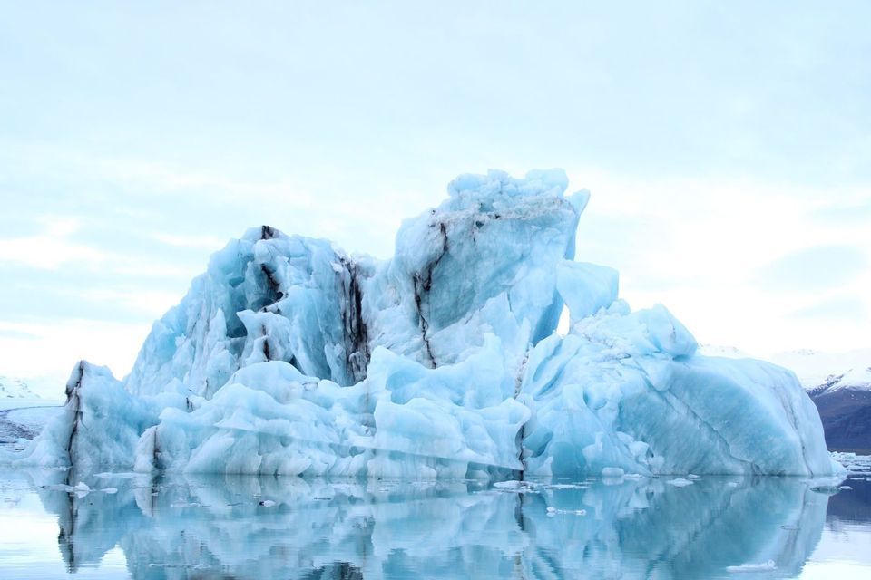 Un enorme iceberg azul dentado flota en aguas tranquilas, reflejándose en la superficie bajo un cielo nublado.