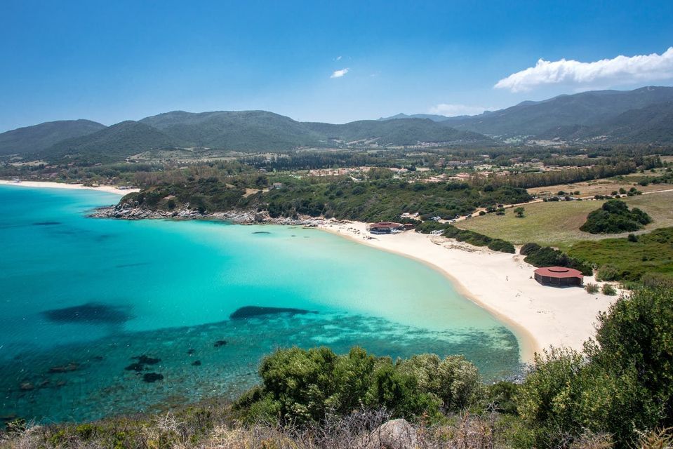 Vista aérea de una playa de arena que se curva por la costa, con aguas turquesas cristalinas y colinas verdes bajo un cielo azul.