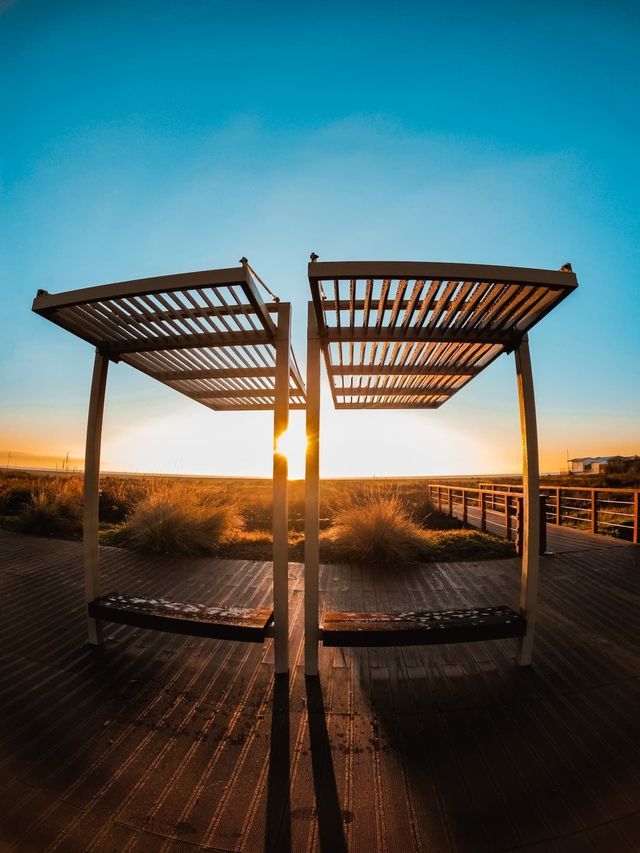Zwei moderne Bänke mit Lamellenüberdachungen auf einem Holzsteg blicken auf eine Graslandschaft während eines goldenen Sonnenuntergangs unter klarem Himmel.