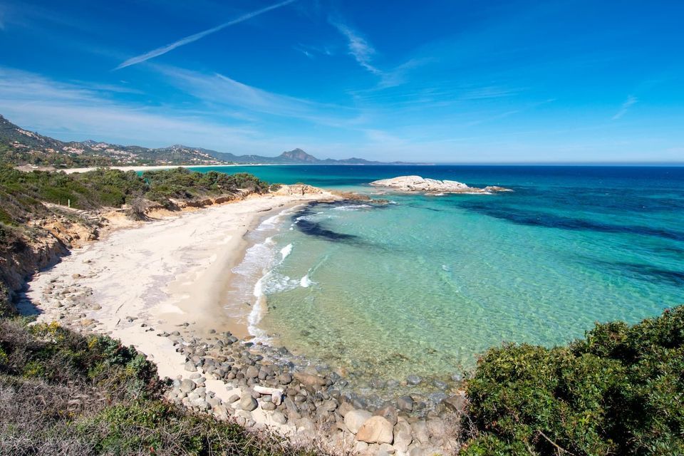 Una vista panorámica de una playa aislada de arena blanca con aguas cristalinas color turquesa, enmarcada por vegetación verde bajo un vasto cielo azul.