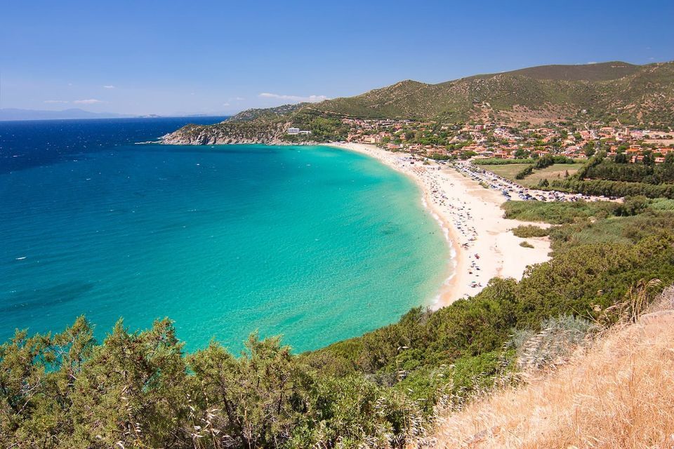Una vista dall'alto di una spiaggia sabbiosa e affollata che si incurva attorno a una baia di acqua turchese, con una cittadina costiera e colline verdi sullo sfondo.
