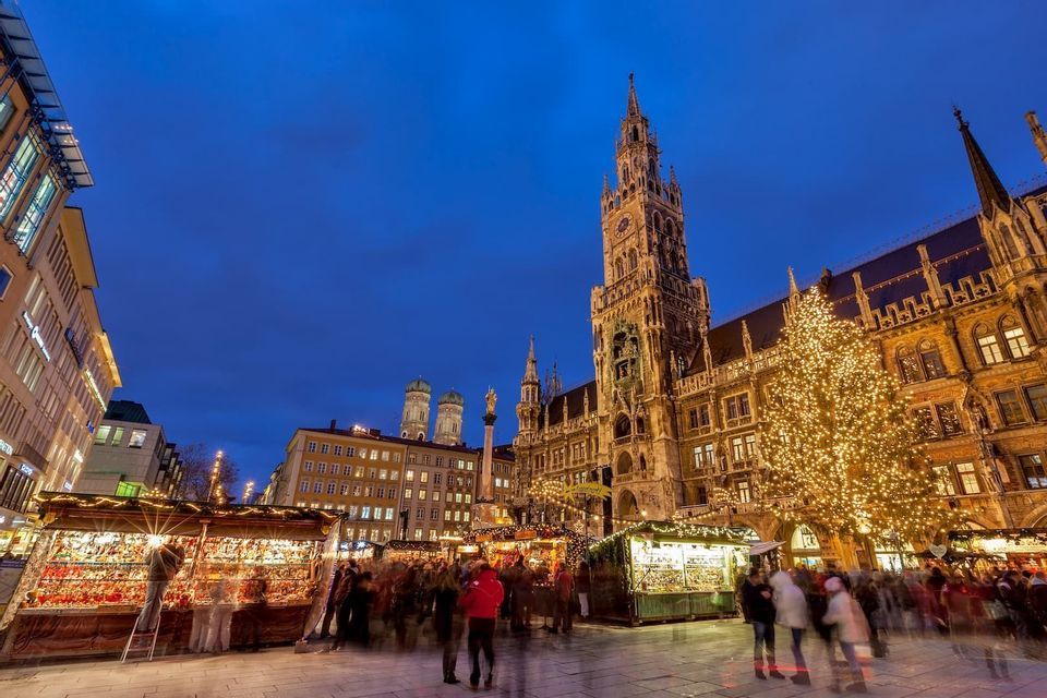 Un animado mercado navideño en una plaza histórica de la ciudad al anochecer, con un árbol iluminado y una ornamentada torre del reloj contra un cielo azul profundo.