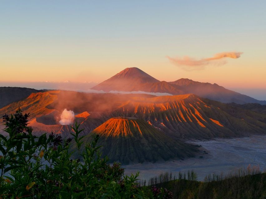 A volcanic landscape with multiple craters, one smoking, is illuminated by the golden light of sunrise against a clear sky.