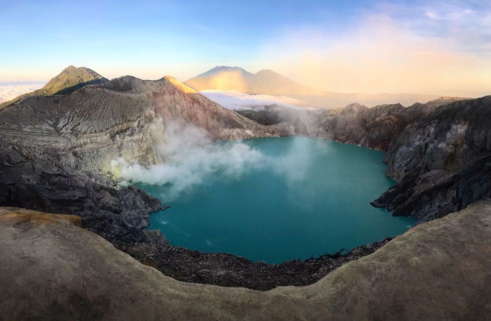 A panoramic view of a turquoise volcanic crater lake with steam rising from its rocky shores and distant mountains visible at sunrise.