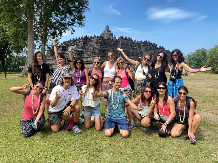 A WeRoad group trip posing and smiling on a grassy lawn in front of a large, ancient stone temple on a sunny day.