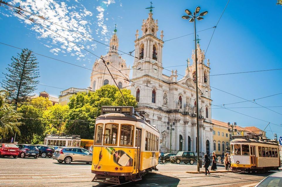 A classic yellow tram on a city street in front of an ornate white basilica under a sunny blue sky with overhead cables.