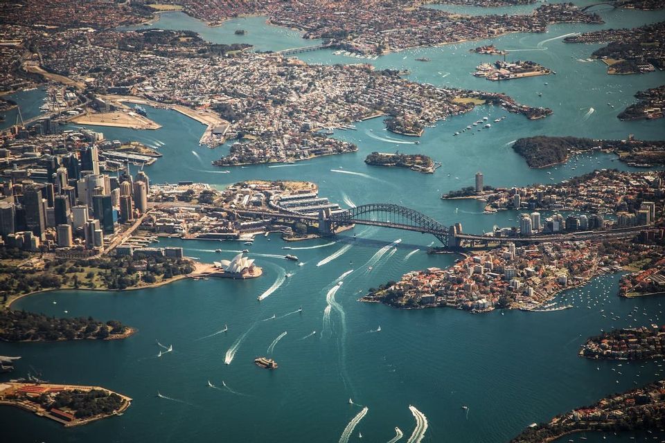 An aerial view of a sprawling city harbor featuring a large arch bridge and an iconic building with a sail-like roof.