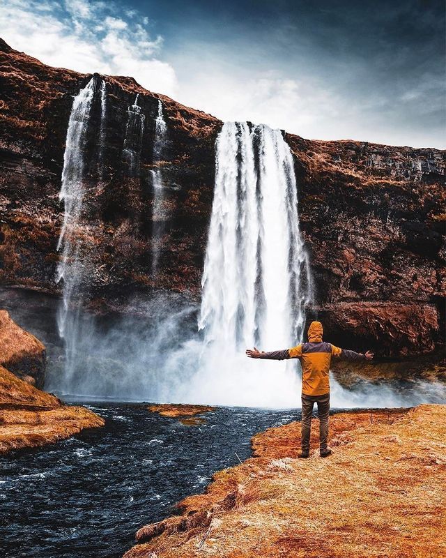 A person in a yellow jacket stands with arms outstretched on a grassy bank, facing a powerful waterfall cascading down a rocky cliff.