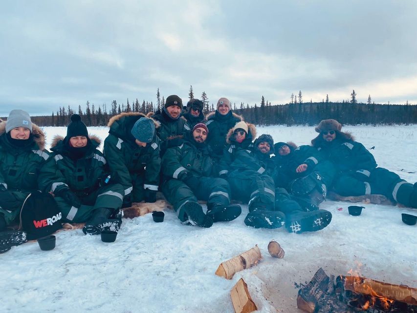 A WeRoad group trip wearing winter gear sits together in the snow around a bonfire, with a forest in the background.