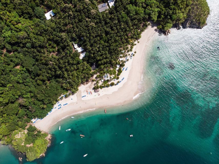 An aerial, top-down view of a tropical white-sand beach cove with turquoise water, small boats, and lush green forest.