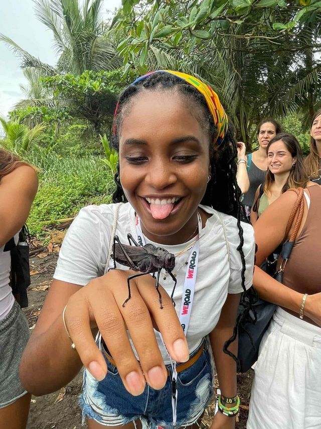 A woman on a WeRoad group trip playfully sticks her tongue out as a large black insect sits on her hand in a jungle.