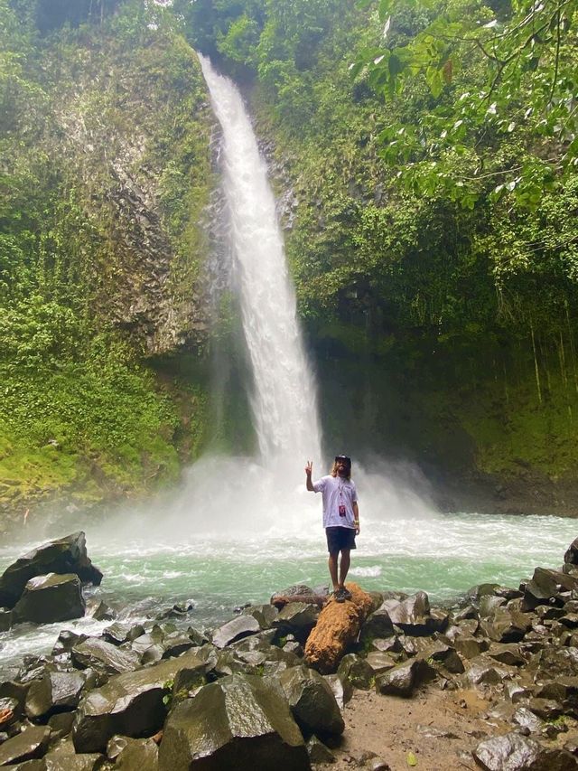A person with long hair stands on a log by a river, making a peace sign with a large waterfall in the background.