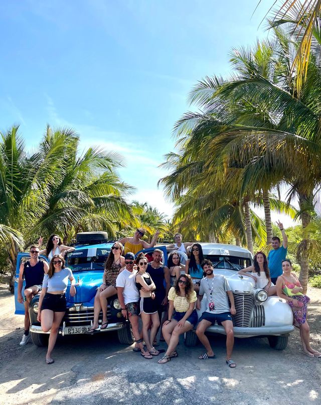 A WeRoad group trip posing for a photo with two vintage cars under tall palm trees on a sunny day.