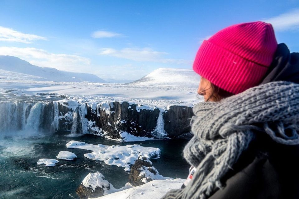 A person in a pink beanie and grey scarf looks out over a large, partially frozen waterfall in a snowy landscape.