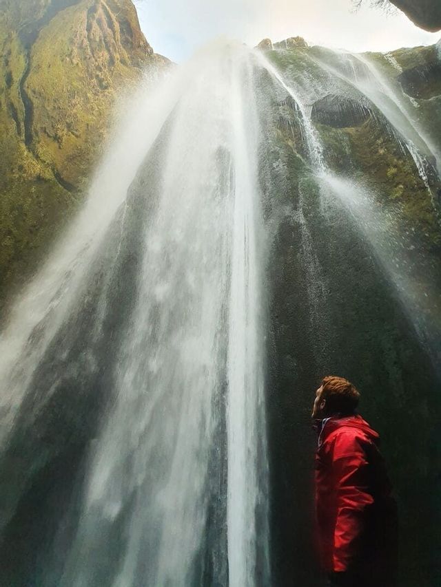 A person wearing a red jacket looks up from below at a tall waterfall cascading down a mossy rock face.