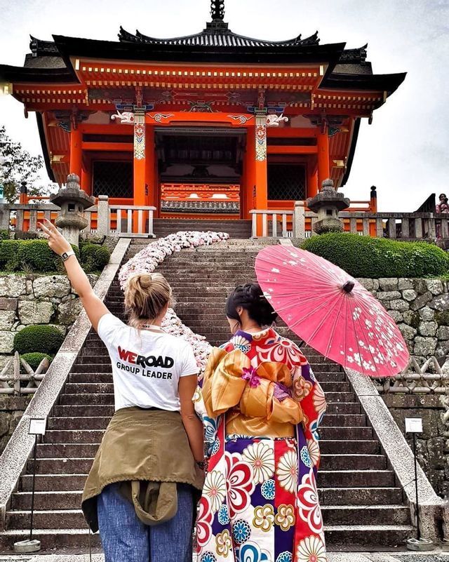 A WeRoad group leader and a woman in a kimono stand on stone steps in front of a traditional orange Japanese temple.