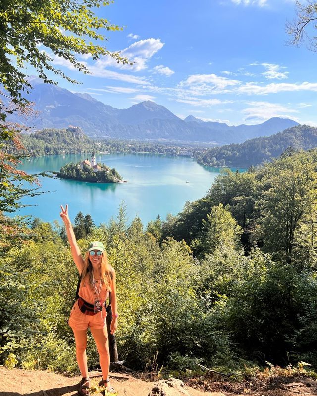 A woman makes a peace sign on a viewpoint above a turquoise lake with an island church, surrounded by forests and mountains.