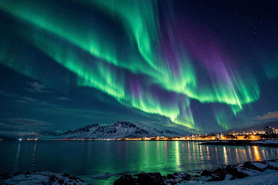 L'aurora boreale verde e viola illumina il cielo notturno stellato sopra una città costiera illuminata e montagne innevate.