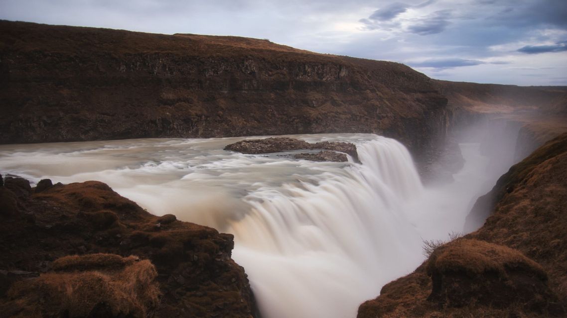 Una fotografia a lunga esposizione di una vasta cascata che precipita in un canyon nebbioso, fiancheggiato da scogliere rocciose marroni sotto un cielo nuvoloso.