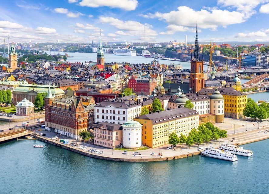 Vista aérea de una ciudad europea histórica con edificios coloridos y agujas a lo largo de un paseo marítimo bajo un cielo azul.