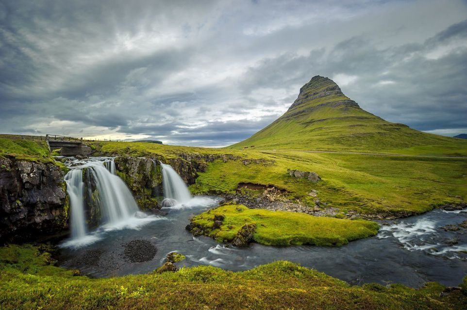 Ein Wasserfall stürzt über Felsen in einen Fluss, mit einem großen, grünen kegelförmigen Berg im Hintergrund unter bewölktem Himmel.