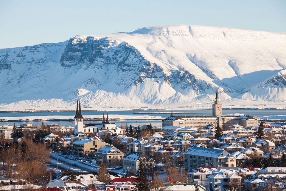 Una veduta aerea di una città innevata con tetti colorati e campanili di chiese, adagiata di fronte a un'imponente montagna innevata oltre uno specchio d'acqua.